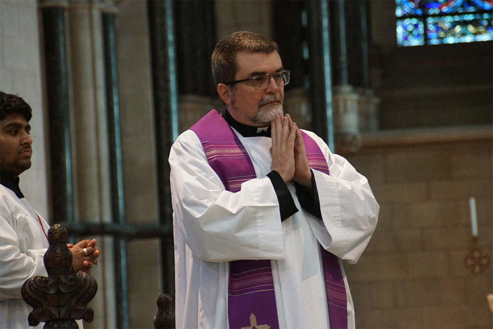 Alan Hodgson Clergy standing with hands in prayer.