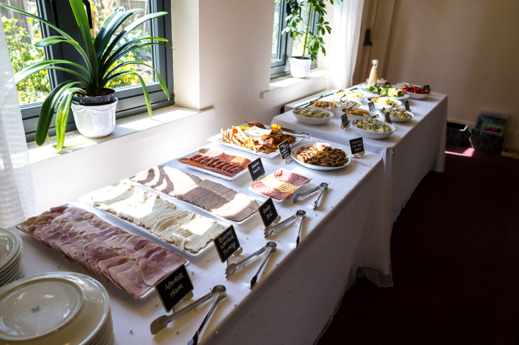 Long table containing wedding buffet food on trays in front of windows.